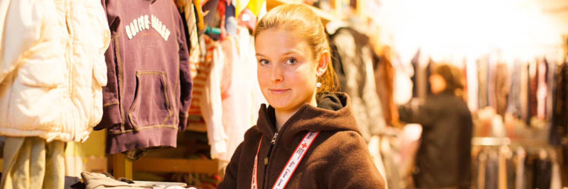 Foto: Eine Mitarbeiterin des Rotkreuz-Shops zeigt einer jungen Frau ein Kleidungsstück mit Kleiderbügel. Die junge Frau freut sich.
