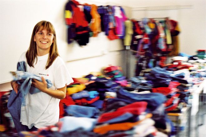 Foto: Eine junge Mitarbeiterin der DRK-Kleiderkammer steht zwischen mehreren Kleiderstapeln. Sie hält eine Jeans in den Händen und blickt lachend in die Kamera.