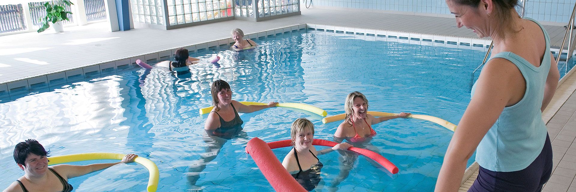 Foto: Mehrere Frauen befinden sich mit Schwimmnudeln in einem Schwimmbecken und blicken die Kursleiterin an. Sie haben sichtbar Spaß zusammen.